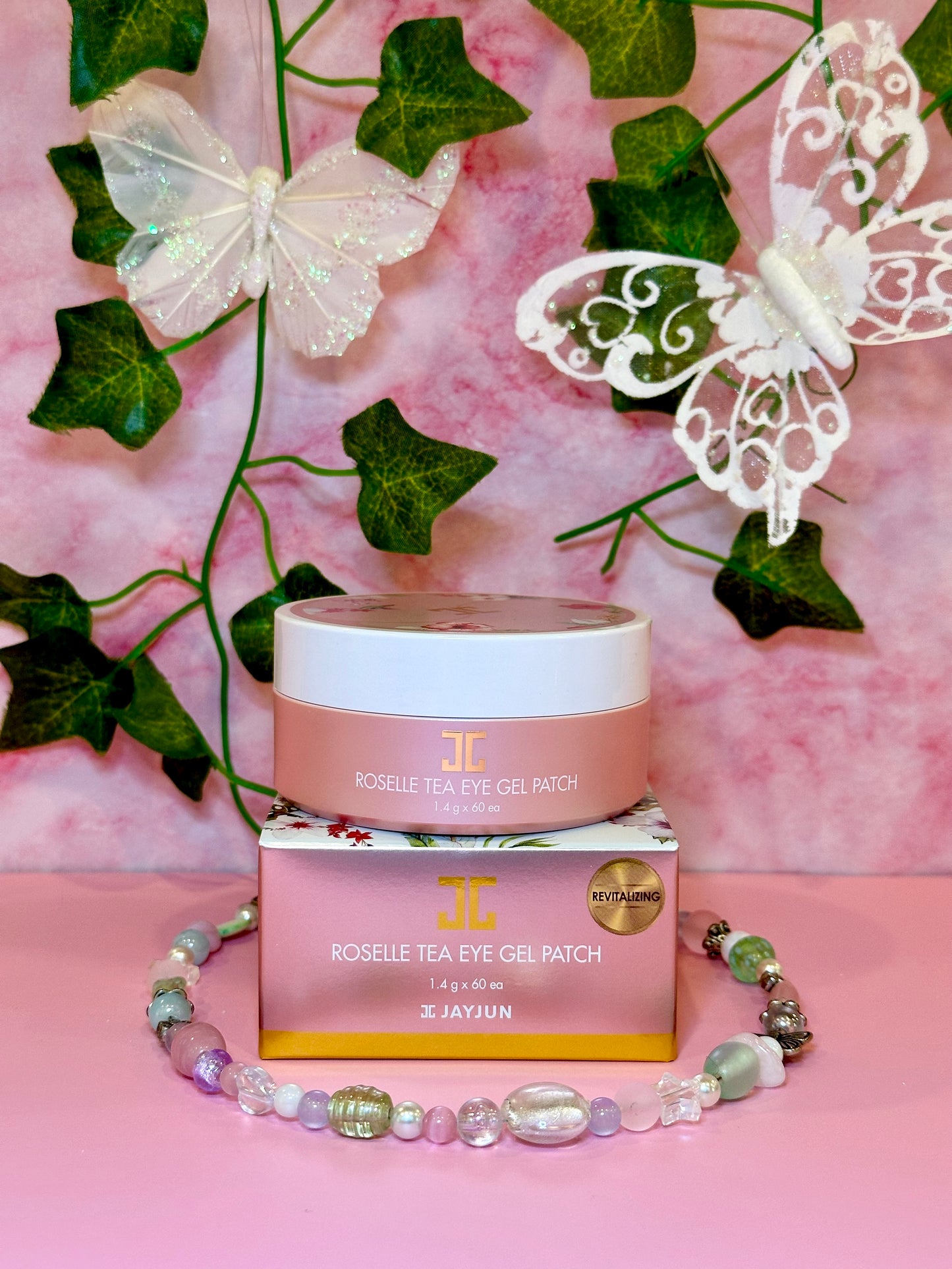The JayJun Roselle Tea Eye Gel Patch jar set against a whimsical backdrop adorned with butterflies and floral decor. This charming presentation highlights the restorative benefits of these Eye Gel Patches.