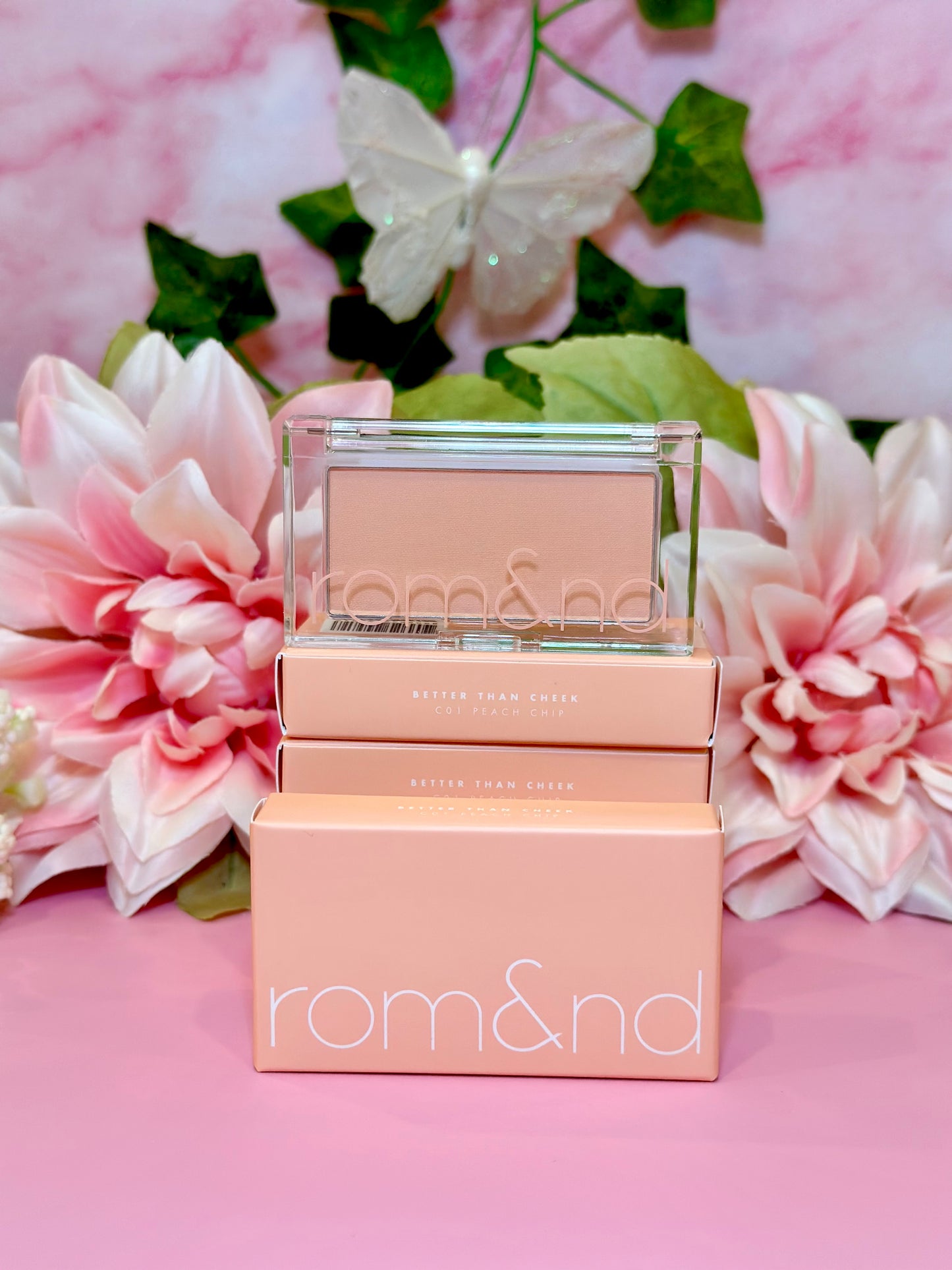 A close-up of the rom&nd Better Than Cheek blush in C01 Peach Chip, surrounded by pink flowers and greenery, exuding charm and elegance, ideal for that perfect cheeky glow.