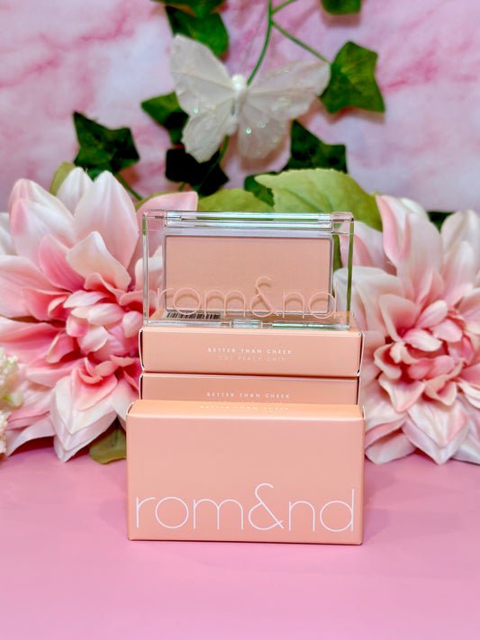 A close-up of the rom&nd Better Than Cheek blush in C01 Peach Chip, surrounded by pink flowers and greenery, exuding charm and elegance, ideal for that perfect cheeky glow.