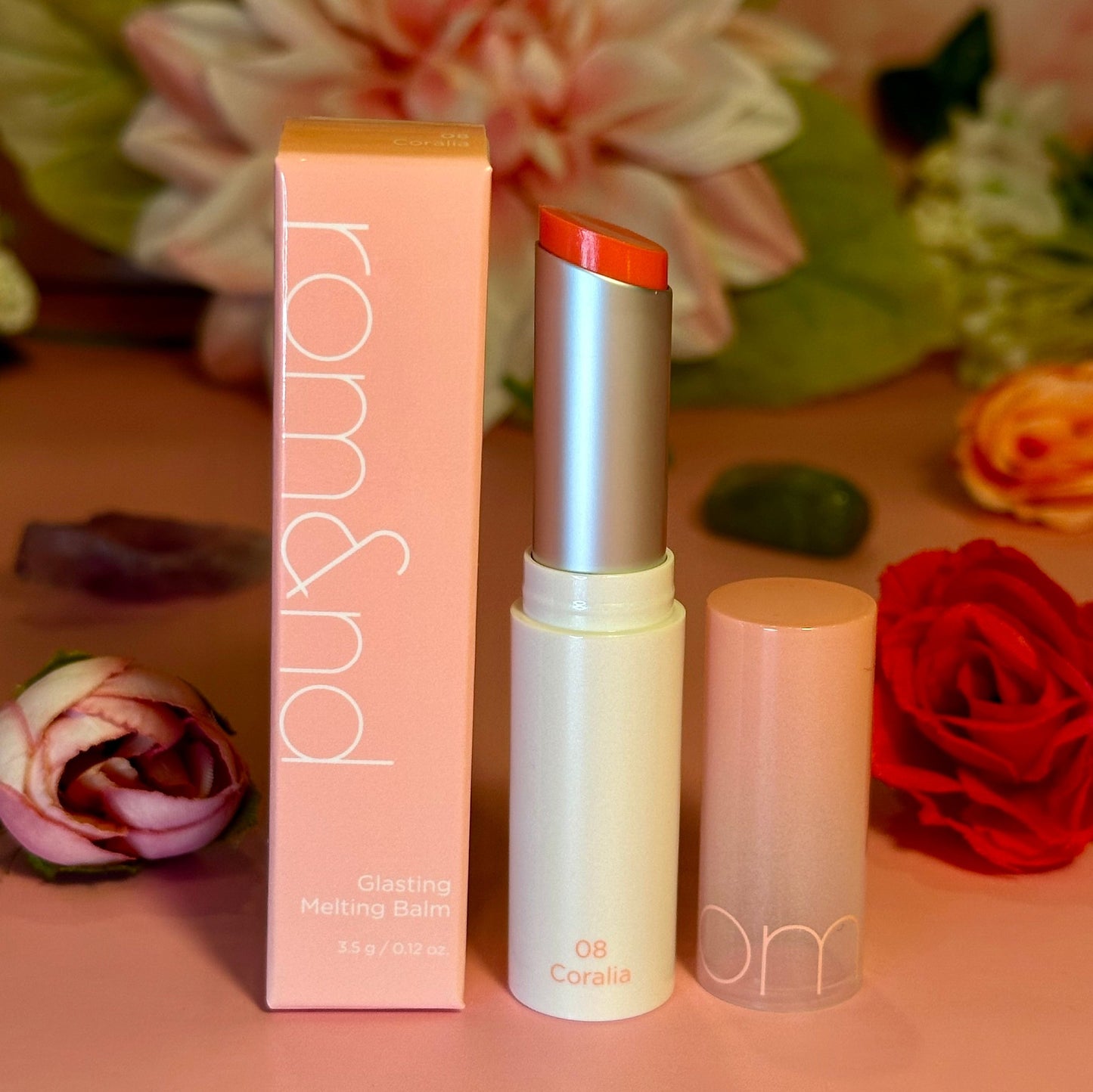 rom&nd Glasting Melting Balm in the shade 08 Coralia, presented with its packaging against a romantic floral backdrop.