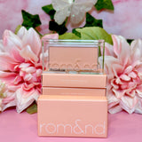 A close-up of the rom&nd Better Than Cheek blush in C01 Peach Chip, surrounded by pink flowers and greenery, exuding charm and elegance, ideal for that perfect cheeky glow.