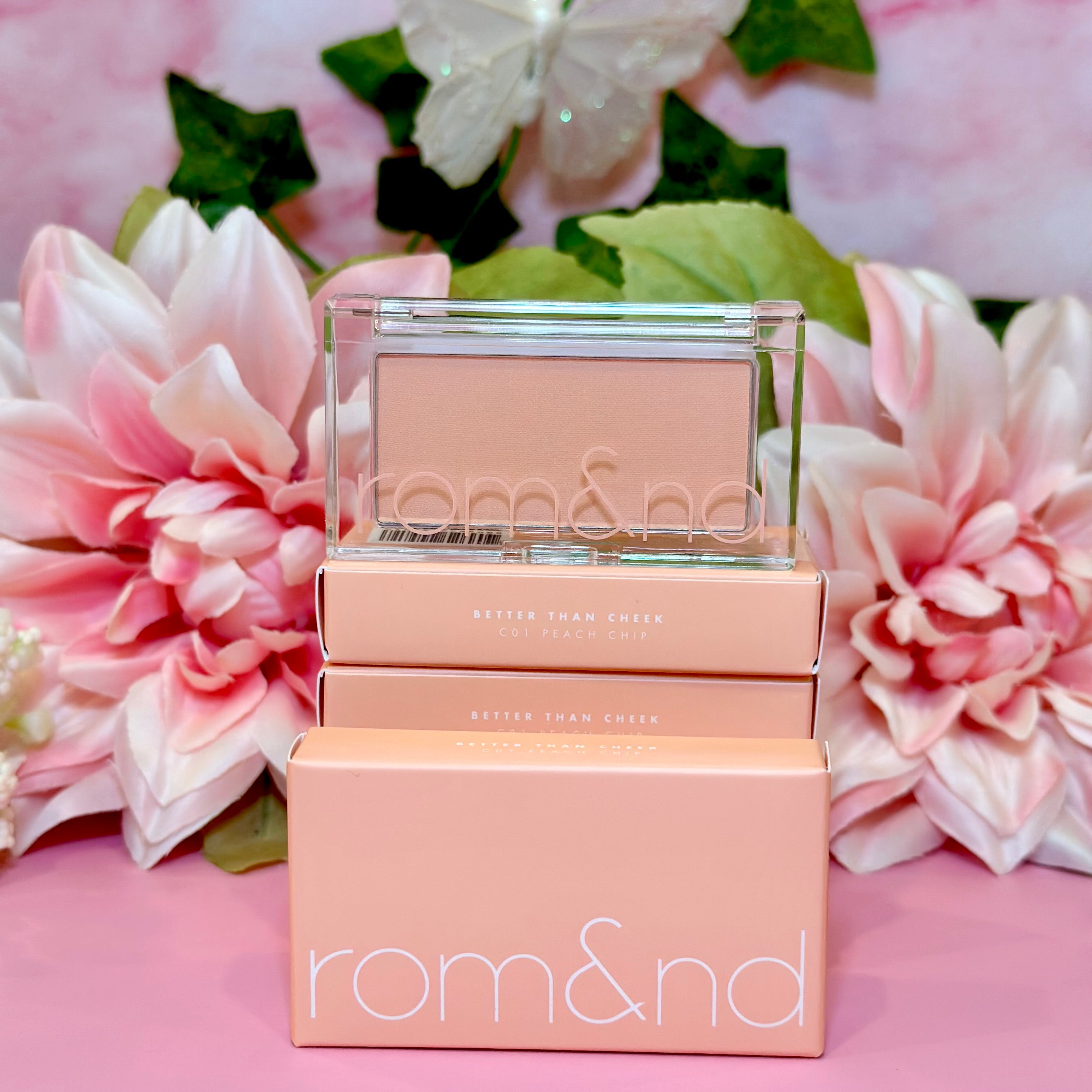 A close-up of the rom&nd Better Than Cheek blush in C01 Peach Chip, surrounded by pink flowers and greenery, exuding charm and elegance, ideal for that perfect cheeky glow.
