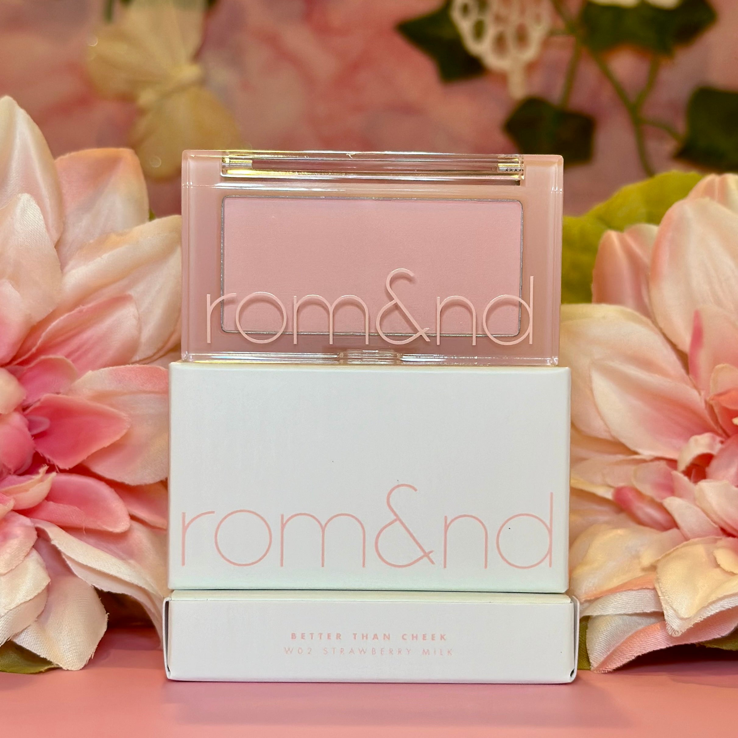 Cheerful presentation of rom&nd Better Than Cheek blush in W02 Strawberry Milk, nestled among pink flowers, radiating soft hues for a fresh makeup look.