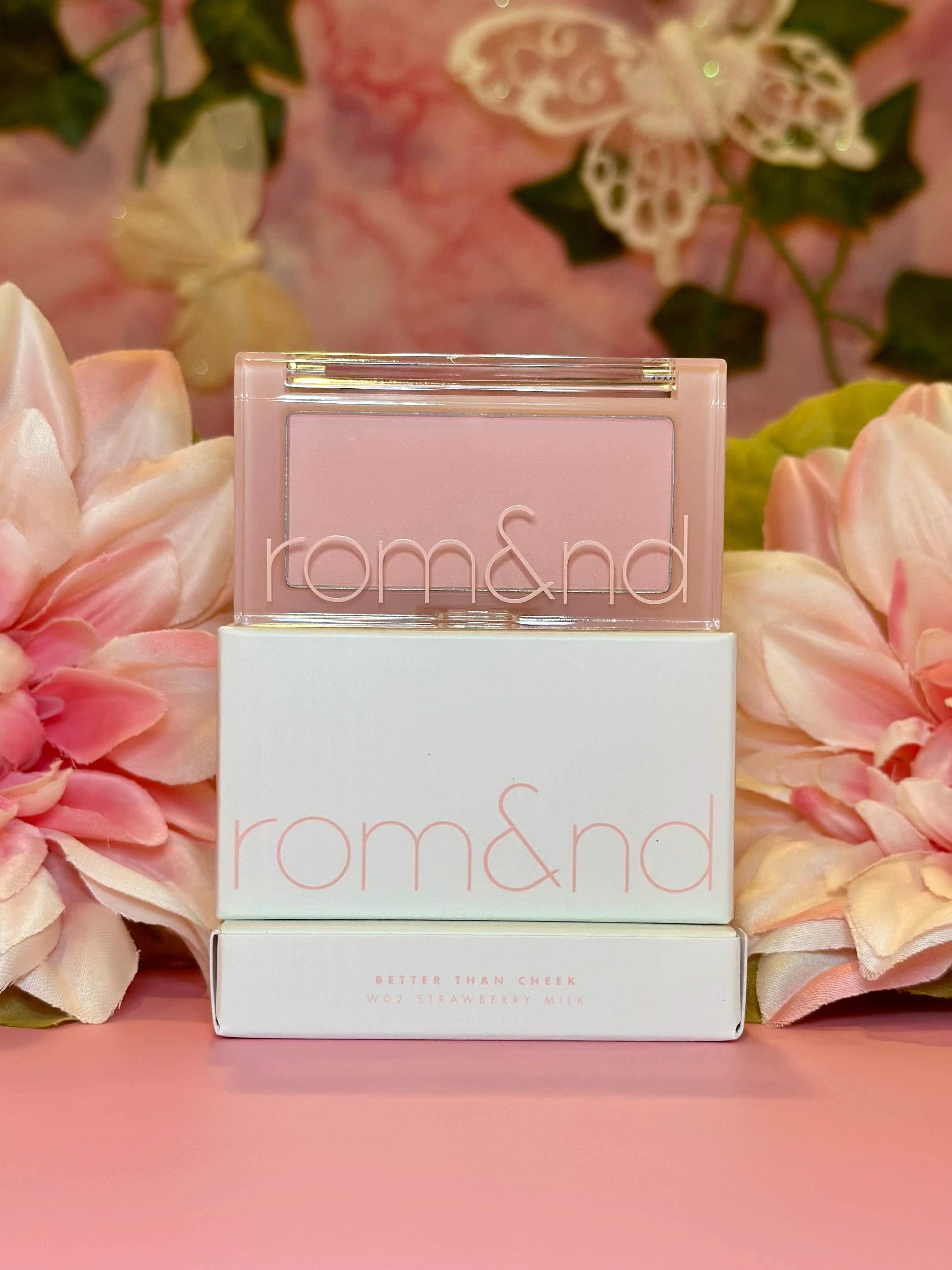 Cheerful presentation of rom&nd Better Than Cheek blush in W02 Strawberry Milk, nestled among pink flowers, radiating soft hues for a fresh makeup look.