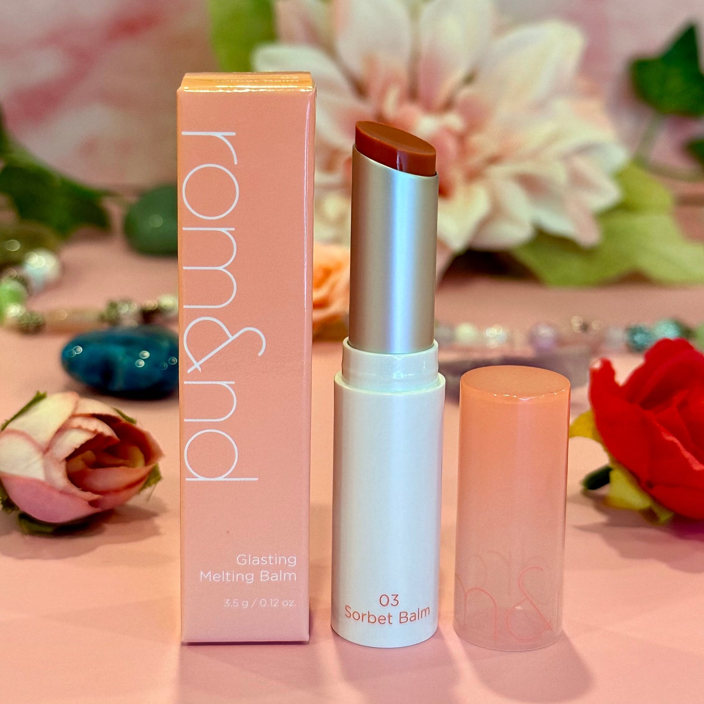 rom&nd Glasting Melting Balm in the shade 03 Sorbet Balm, positioned alongside its pink packaging surrounded by floral elements.